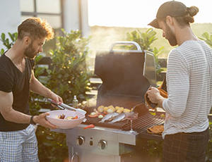 Zwei Männer stehen an einem Gasgrill und haben Grillgut auf dem Grill