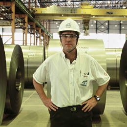 Employé sur un site industriel debout au milieu d'énormes rouleaux d'acier. Il porte un casque et une chemise blanche